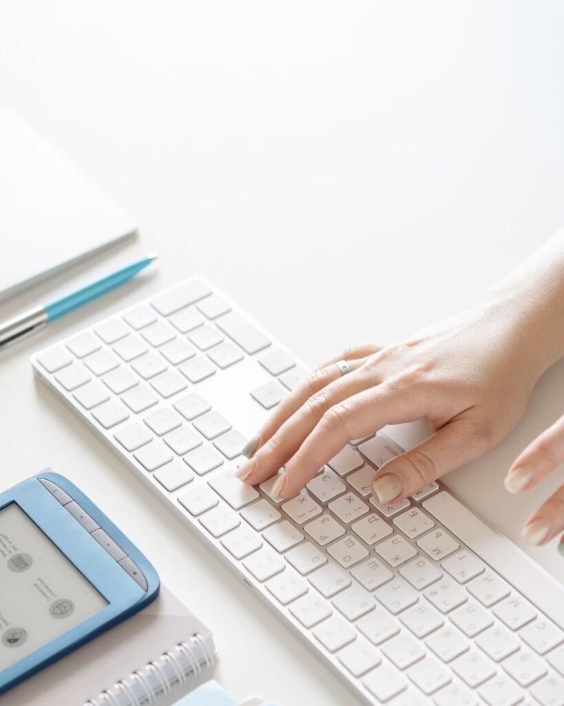 A hand typing on a slim white wireless keyboard on a bright desk beside a blue e-reader and a notebook