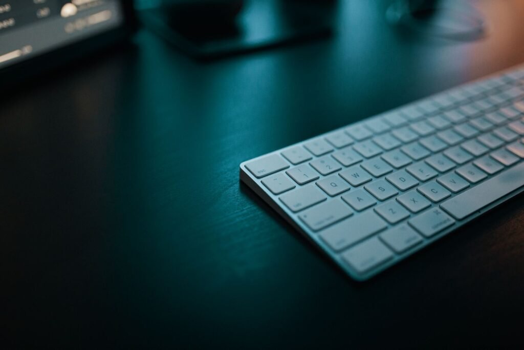 Angled close-up of a slim white low-profile wireless keyboard on a dark wooden desk with a monitor blurred behind it