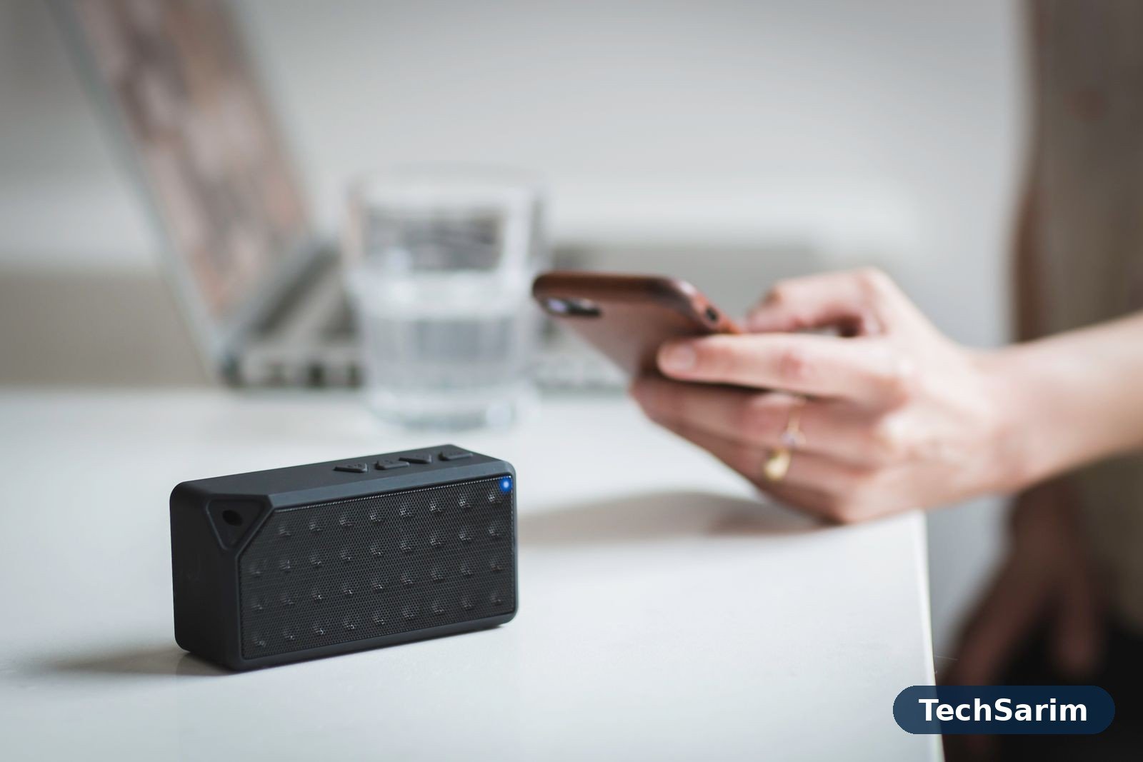 A small black Bluetooth speaker in sharp focus on a white desk while a person holds a smartphone behind it, a laptop blurred in the background