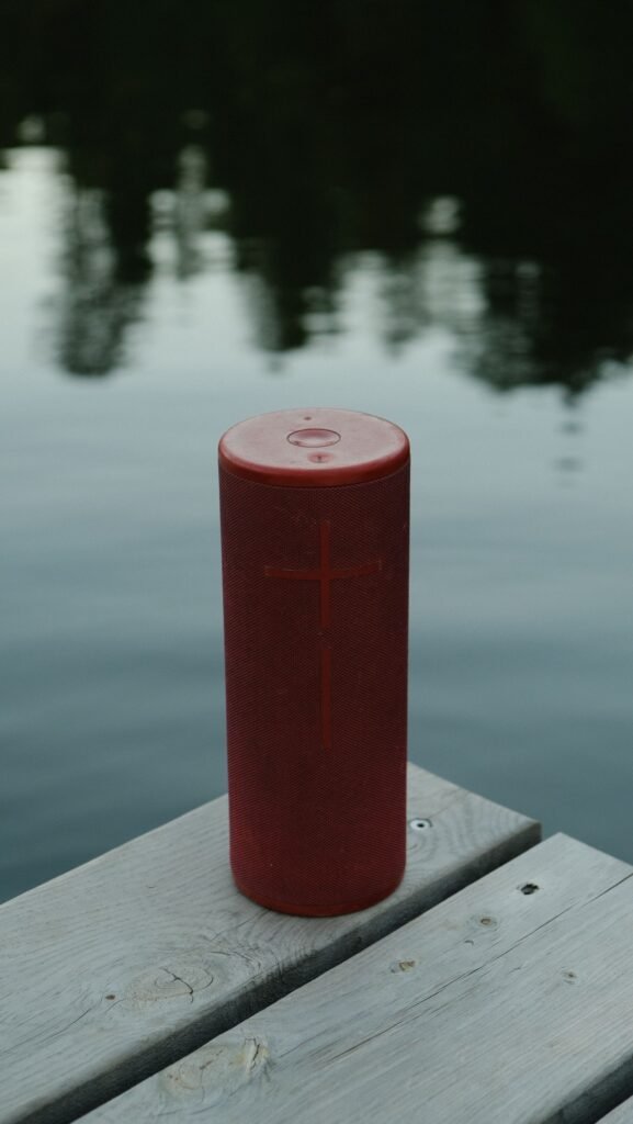 A tall red cylindrical portable speaker standing on a wooden dock beside a calm lake