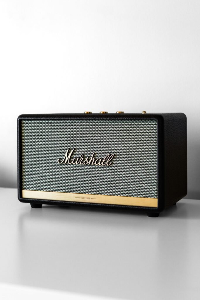 A black Marshall home speaker with a woven grille and gold script logo sitting on a white shelf