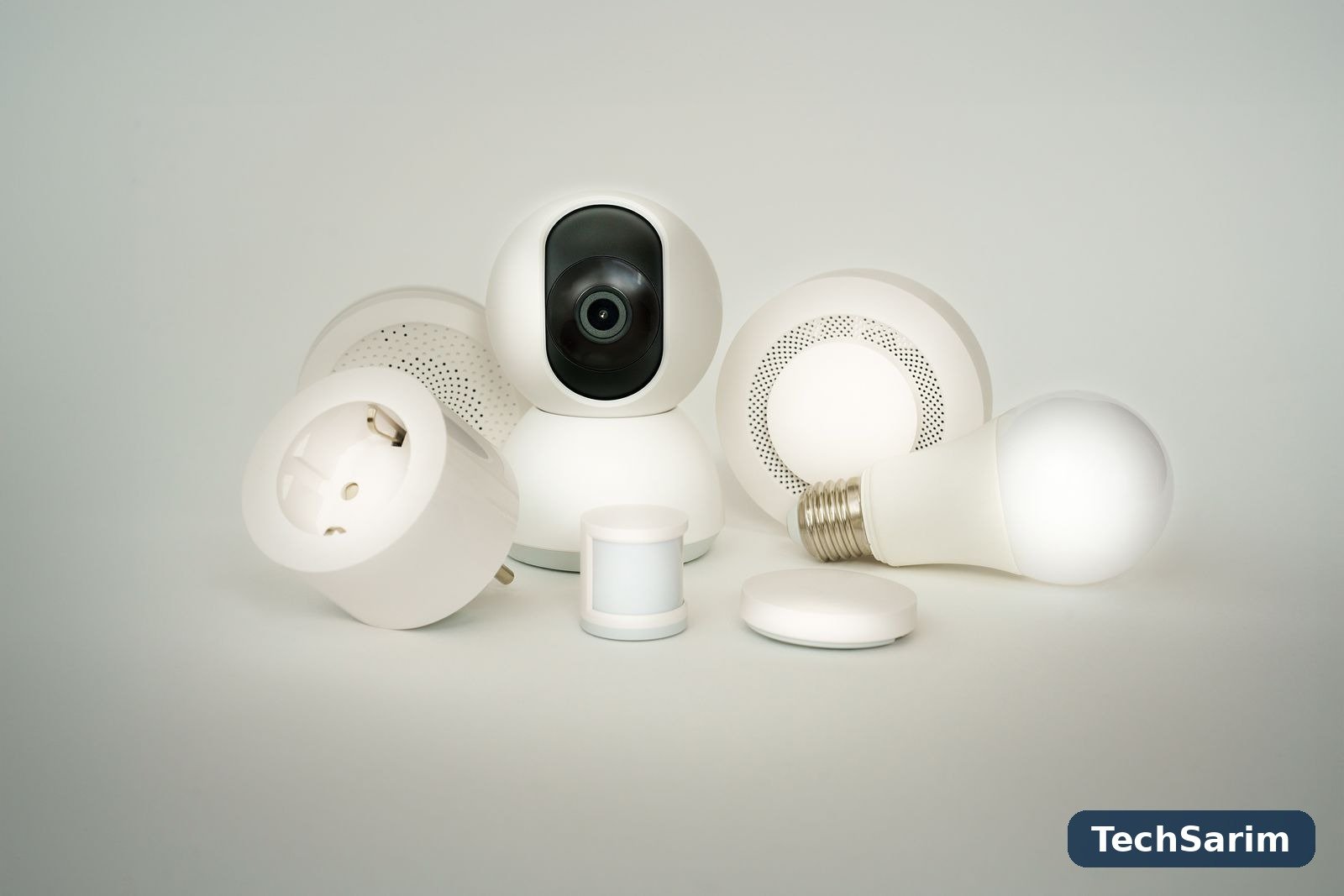 A collection of white smart home devices on a pale background: a pan-and-tilt security camera, a smart plug, a smart bulb, a motion sensor and a round smoke alarm