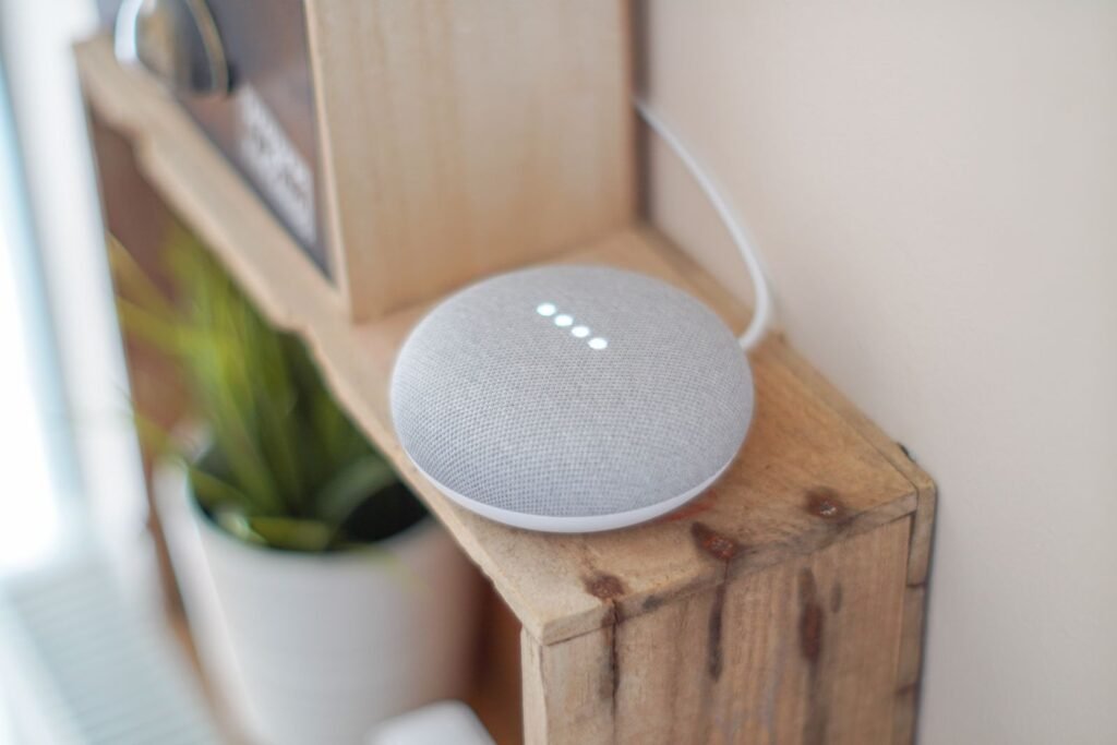 A grey fabric Google Home Mini smart speaker with its four indicator lights glowing, sitting on a rustic wooden shelf next to a potted plant