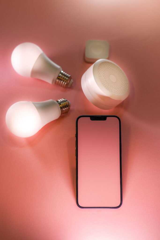 Two glowing white smart light bulbs, a small round smart speaker and a smartphone with a blank screen lying on a pink background