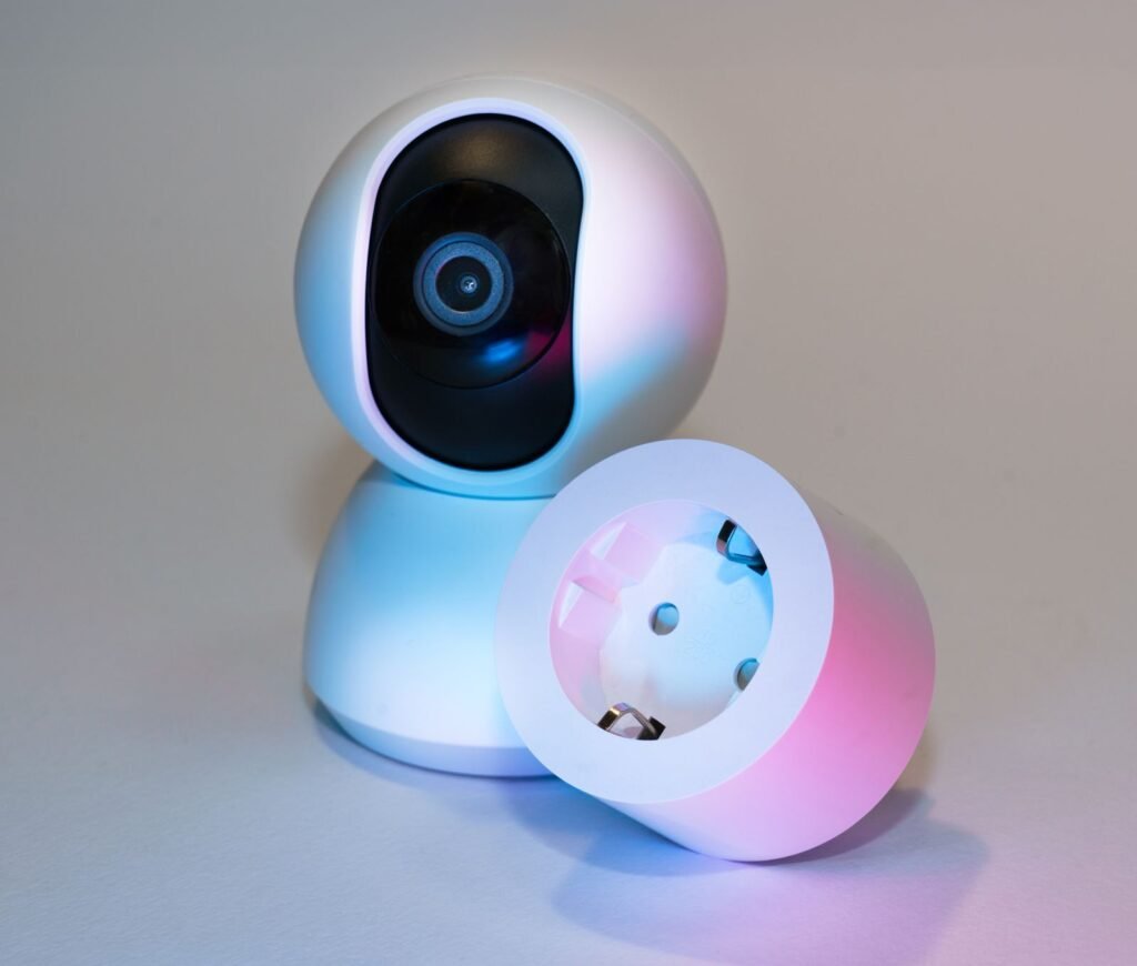 A white pan-and-tilt home security camera standing behind a round white smart plug, lit by blue light on one side and pink on the other