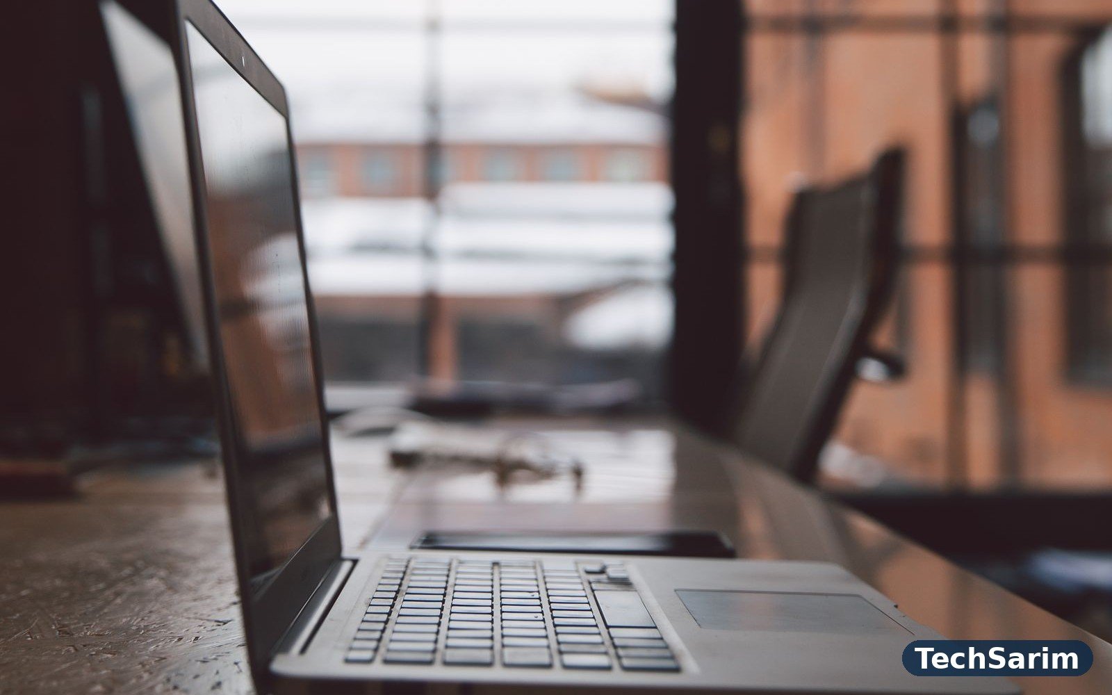 A slim silver laptop open on a long wooden desk in a bright loft office with tall industrial windows