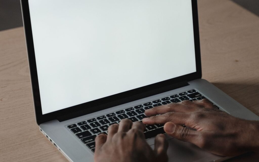 Close-up of hands typing on a silver laptop with a bright blank screen on a light wooden desk