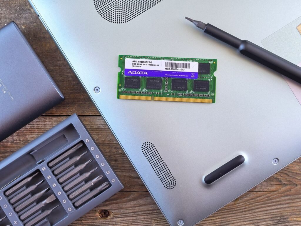 A green laptop RAM stick resting on the underside of a silver laptop beside a precision screwdriver and bit set on a wooden table