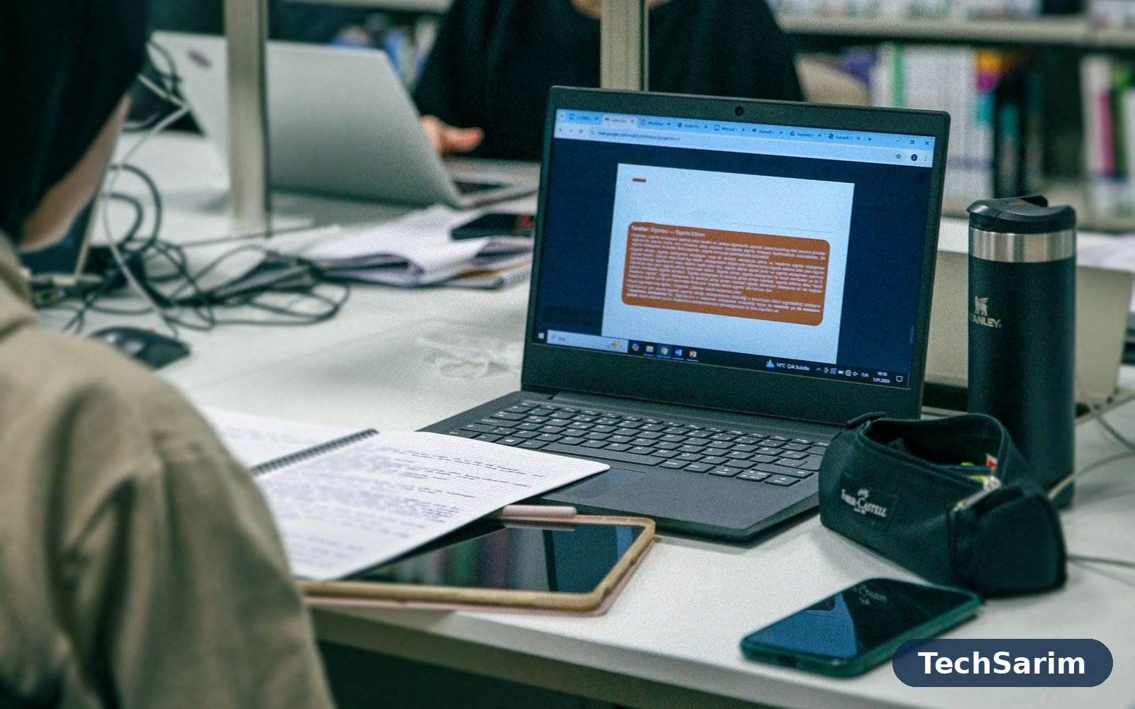A student's library desk with an open laptop showing study slides, a tablet with a stylus, a smartphone, a handwritten notebook and a travel mug