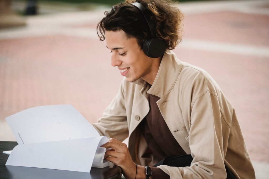 A smiling student wearing black over-ear wireless headphones while reading study papers outdoors