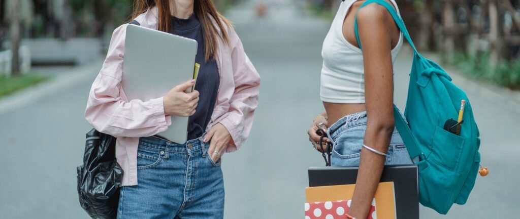 Two students walking outdoors between classes, one carrying a laptop under her arm and the other wearing a teal backpack and holding folders